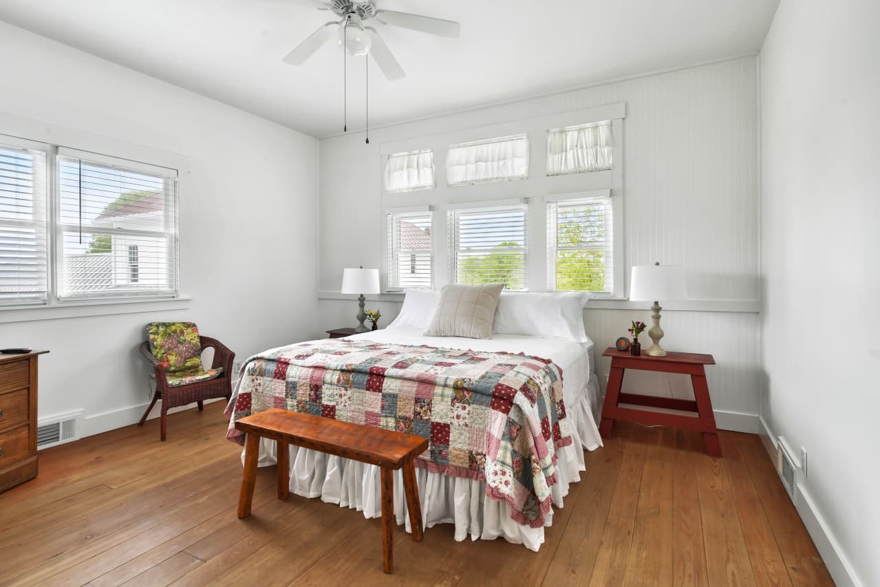 A bright bedroom with wood floors, featuring a white bed with a colorful patchwork quilt, high ceilings, and large windows overlooking trees.
