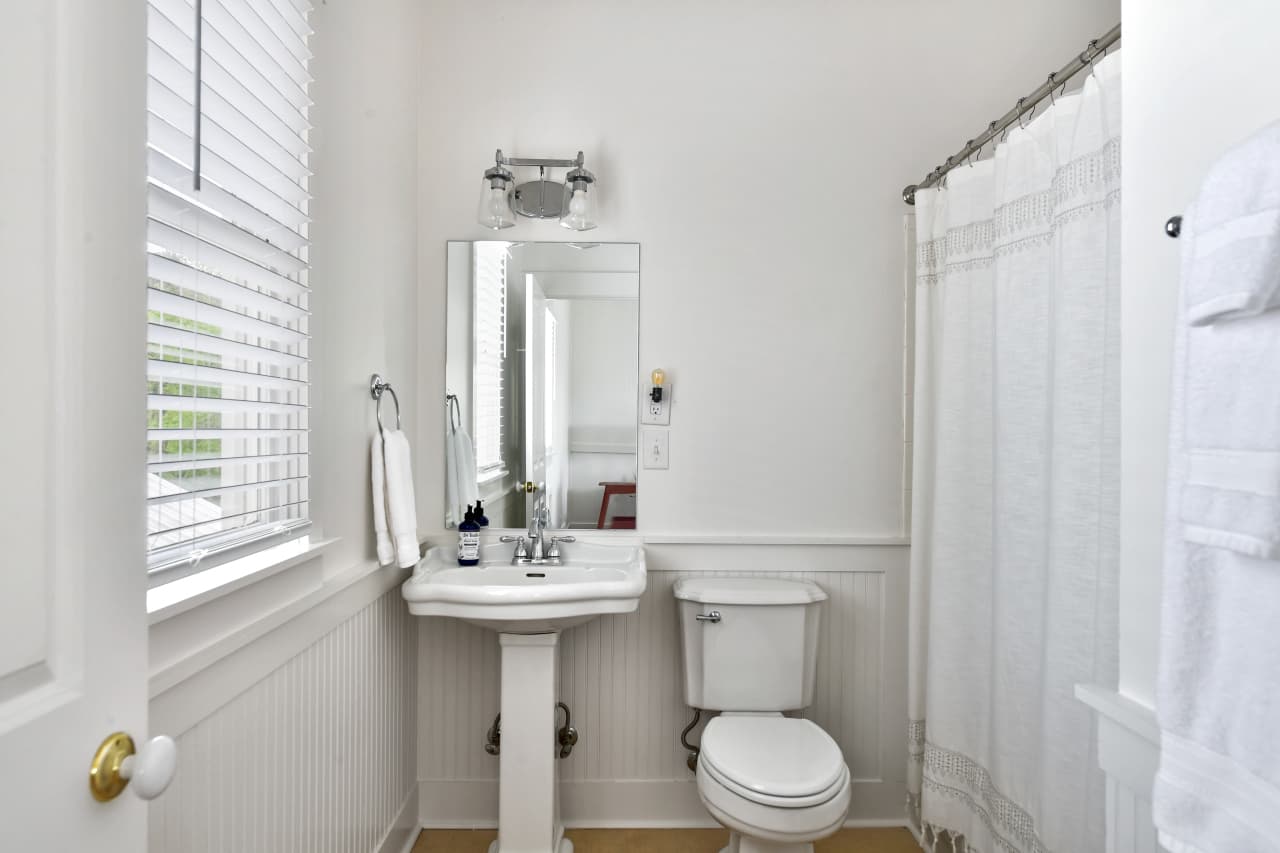 A bright, clean white bathroom featuring a pedestal sink, a toilet, and a shower with a white curtain. A window with blinds lets in natural light.