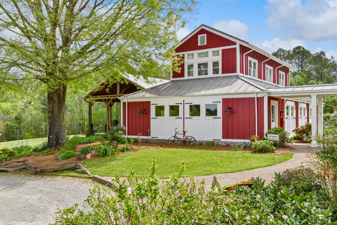 A charming two-story red and white converted barn featuring a metal roof, surrounded by a lush green lawn, mature trees, and a winding stone pathway.