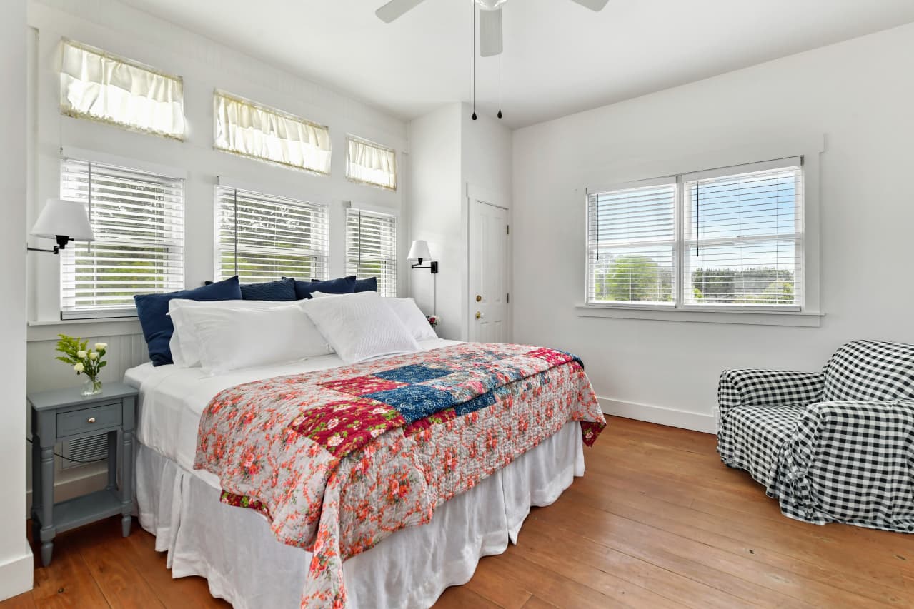 A bright bedroom with wood floors featuring a bed with a vibrant floral and patchwork quilt, a blue nightstand, and a black-and-white checkered armchair near a window.