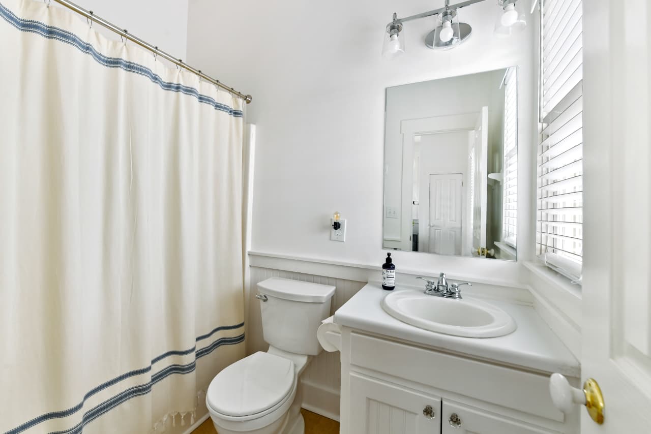 A clean, bright bathroom with a white vanity and sink, a toilet, and a cream-colored shower curtain with blue stripes. A large mirror and vanity light brighten the space.