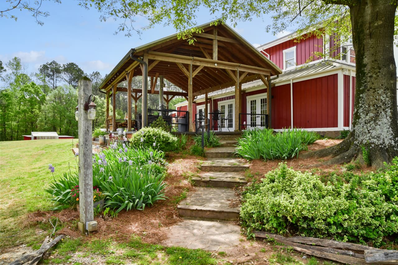 A stone pathway leads through lush gardens and flowering plants toward a large, rustic timber-frame covered porch attached to a red barn-style building.