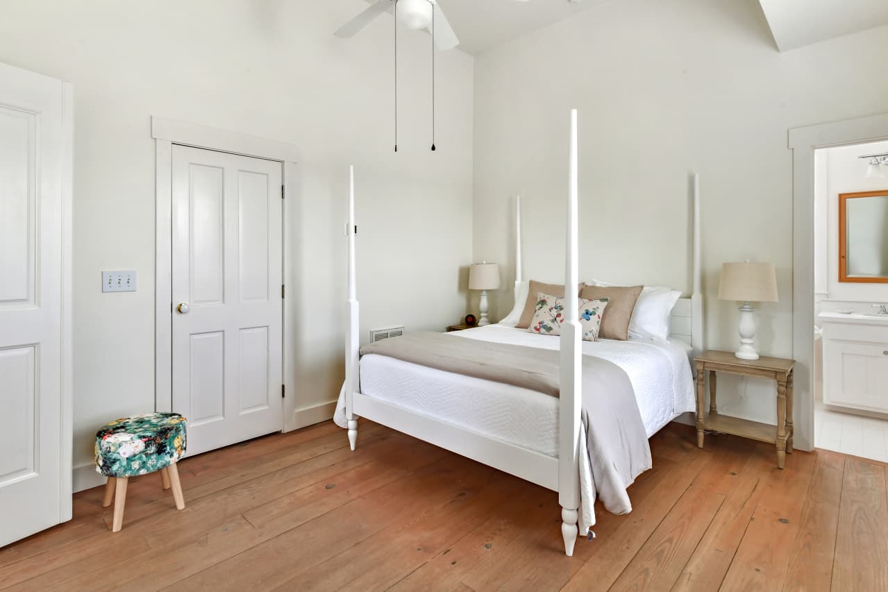 A bright bedroom with high ceilings and wood floors, featuring a white four-poster bed, two bedside lamps, and a glimpse into an en suite bathroom.