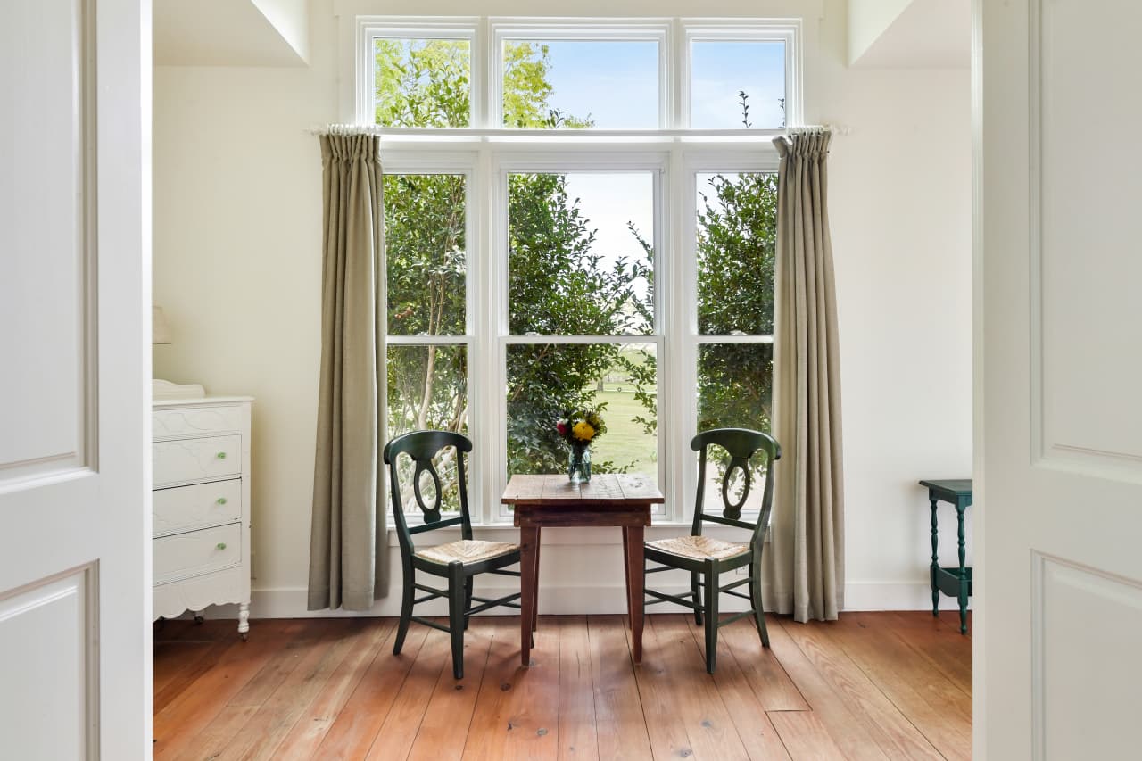 A sunlit sitting area with wood floors, featuring a small wooden table and two dark green chairs positioned in front of a large three-pane window with views of lush greenery.