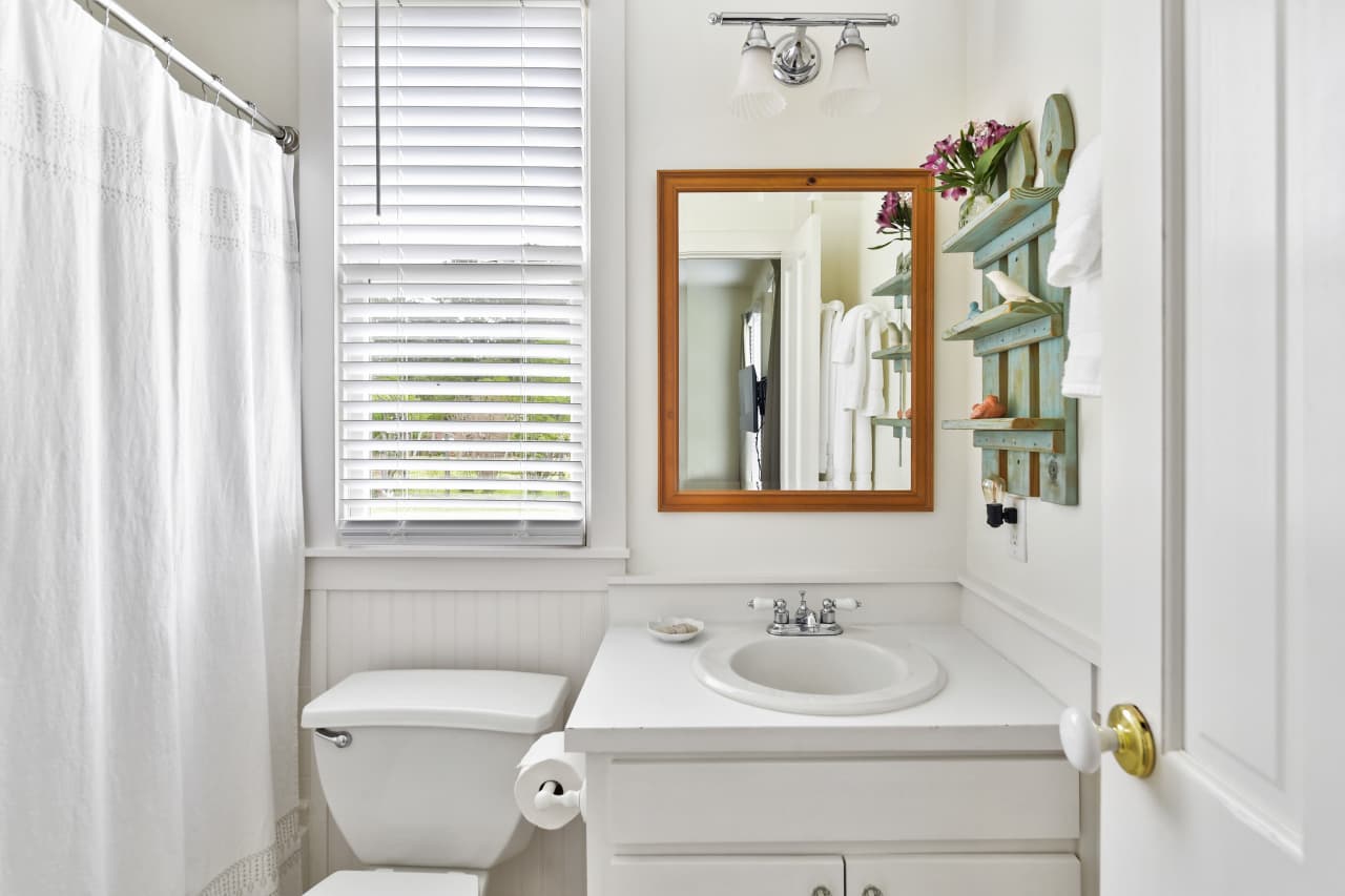 A bright bathroom featuring a white vanity with a framed wood mirror, a toilet, and a white shower curtain. A window with blinds and a small teal wall shelf add light and character.