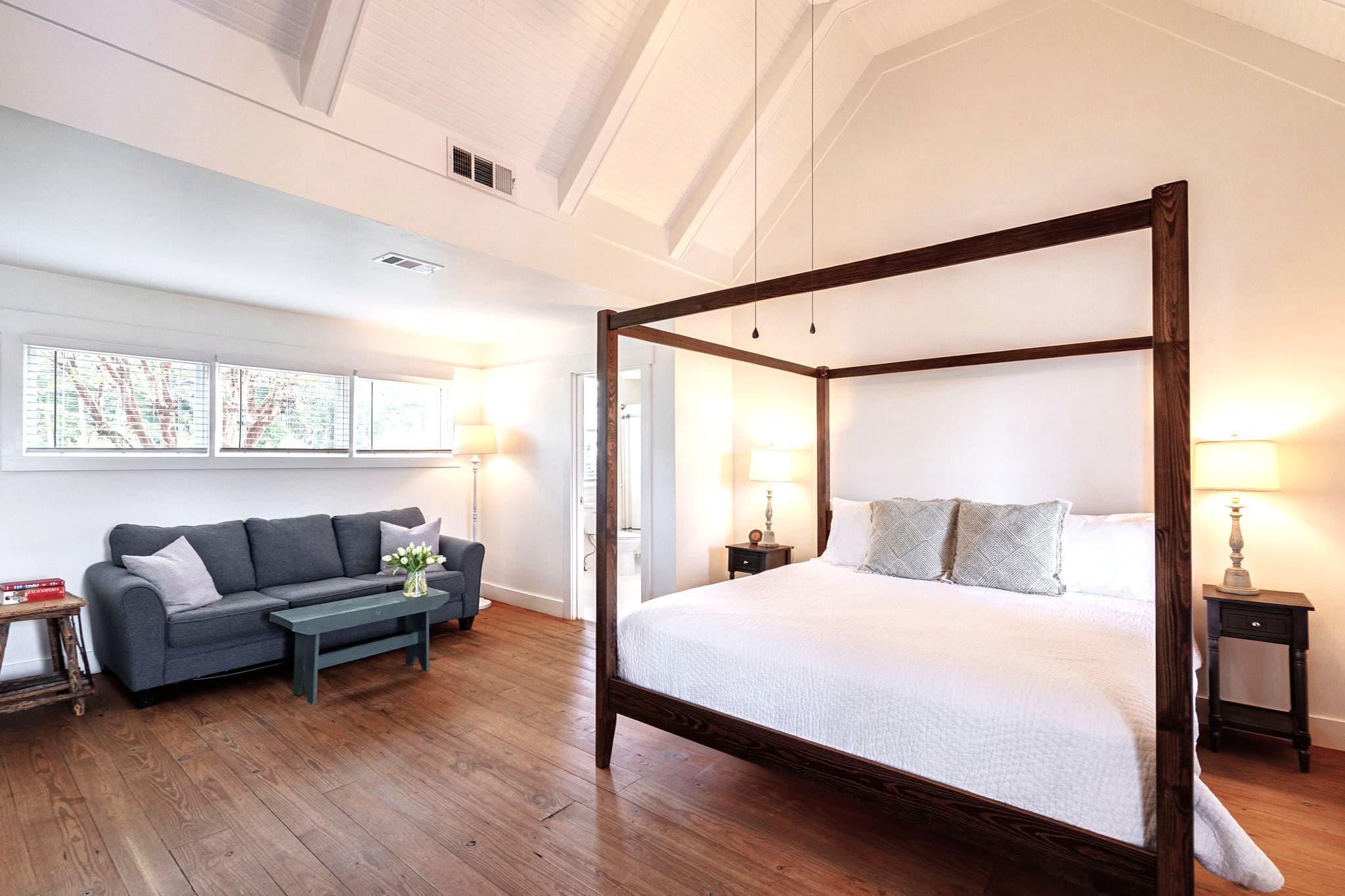 A spacious bedroom with high vaulted ceilings and wood floors, featuring a dark wood four-poster king bed and a separate sitting area with a blue sofa and coffee table.