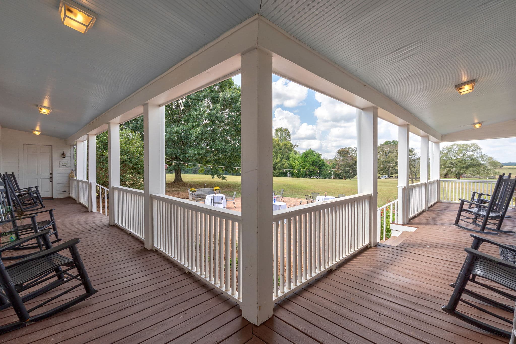 A wide, wrap-around covered porch with dark wood decking and white railings, lined with several black rocking chairs overlooking a grassy field and trees.
