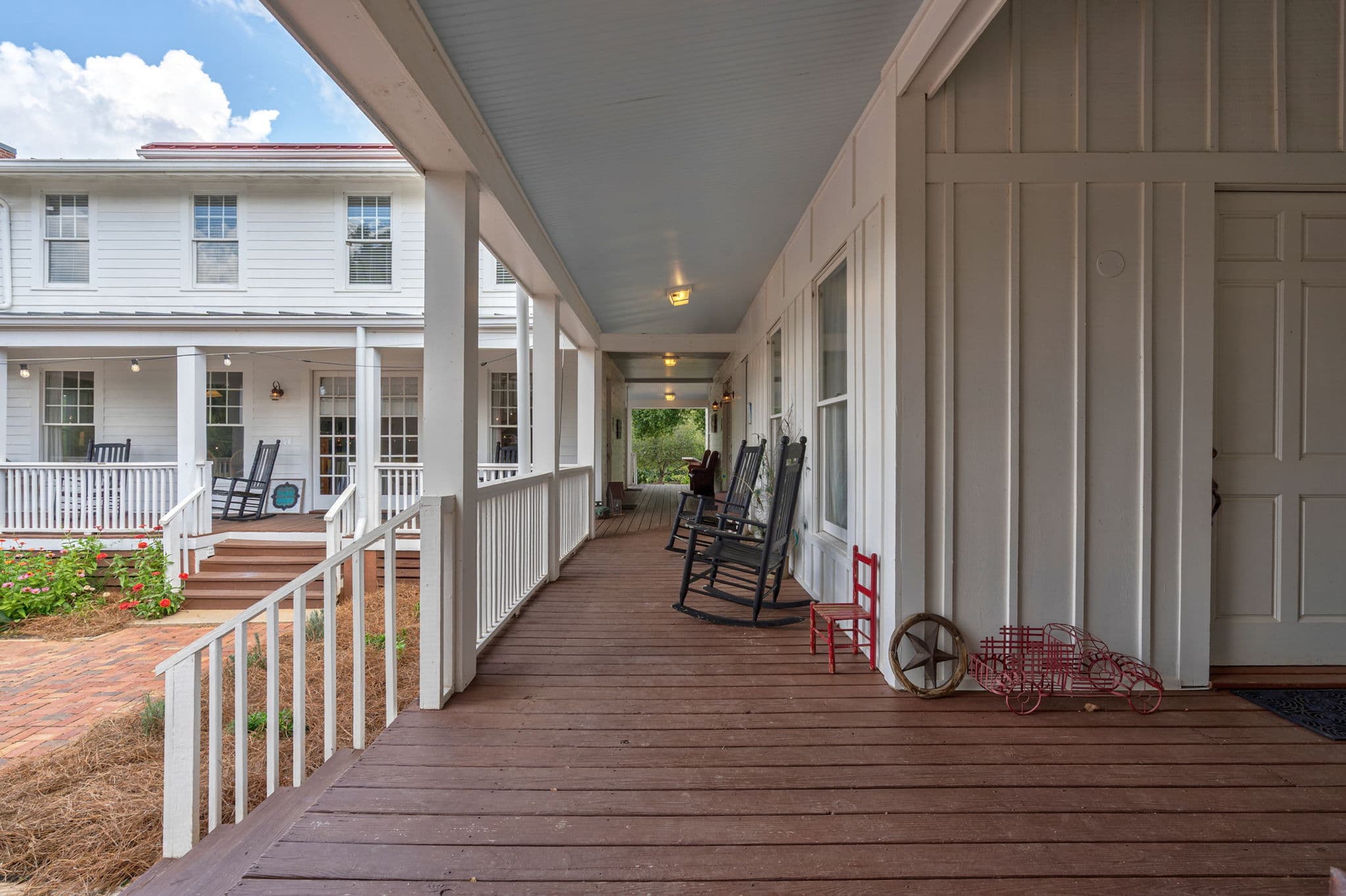 A long, covered white porch with dark wood floors and black rocking chairs, looking toward a large white farmhouse across a courtyard path.