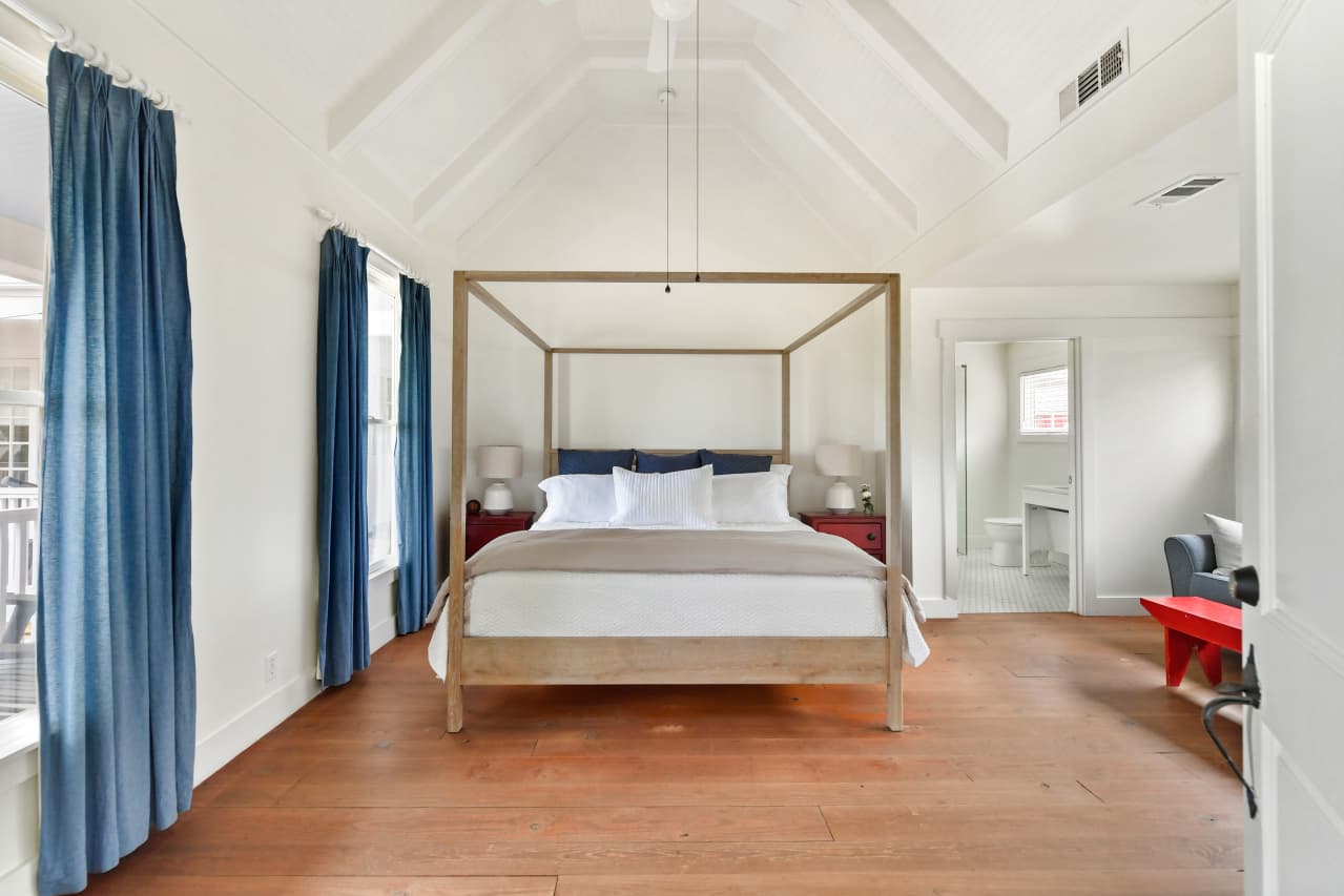 A bright bedroom with soaring vaulted ceilings and wood floors, featuring a light wood four-poster bed framed by blue curtains. A doorway offers a view into an en suite bathroom.