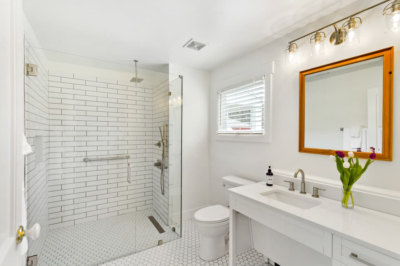 A bright, modern accessible bathroom featuring a large roll-in shower with white subway tiles and a glass partition. It includes a white vanity with a wood-framed mirror and a toilet.