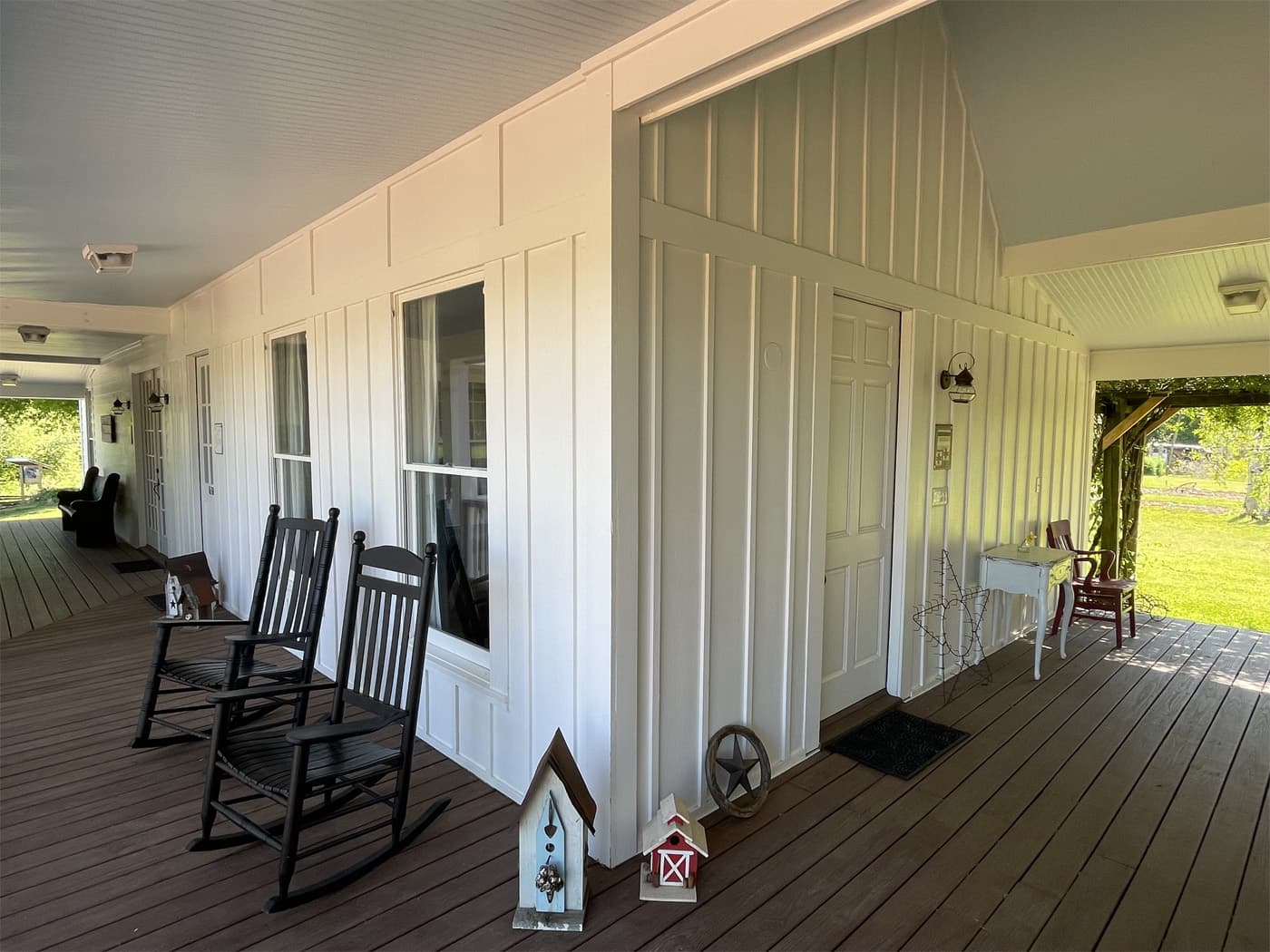 A covered porch with white board-and-batten siding and dark wood floors, featuring black rocking chairs, a private entrance door, and decorative birdhouses.