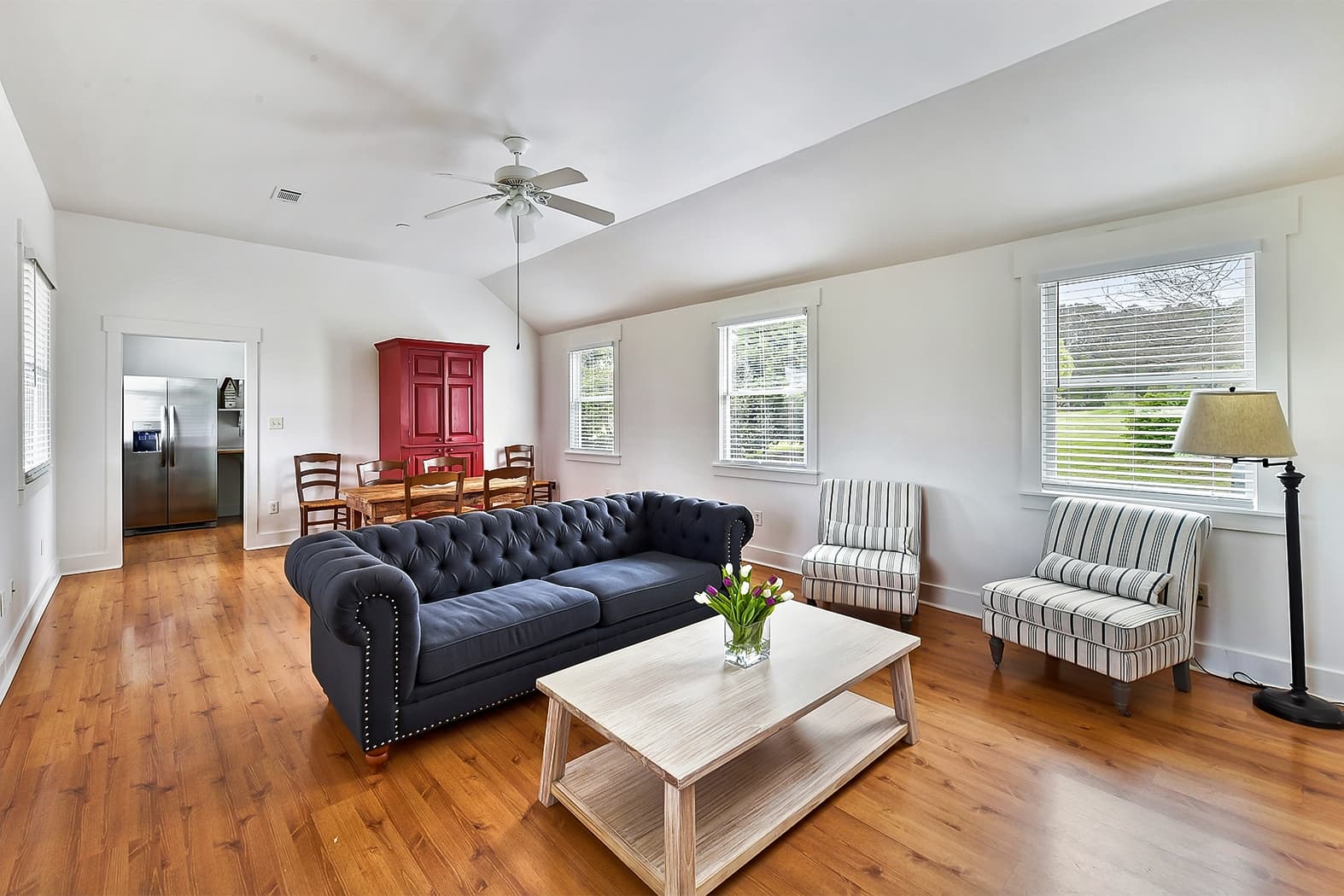A spacious, light-filled living room with wood floors and vaulted ceilings, featuring a navy blue tufted sofa, a light wood coffee table, and several windows overlooking a green landscape.