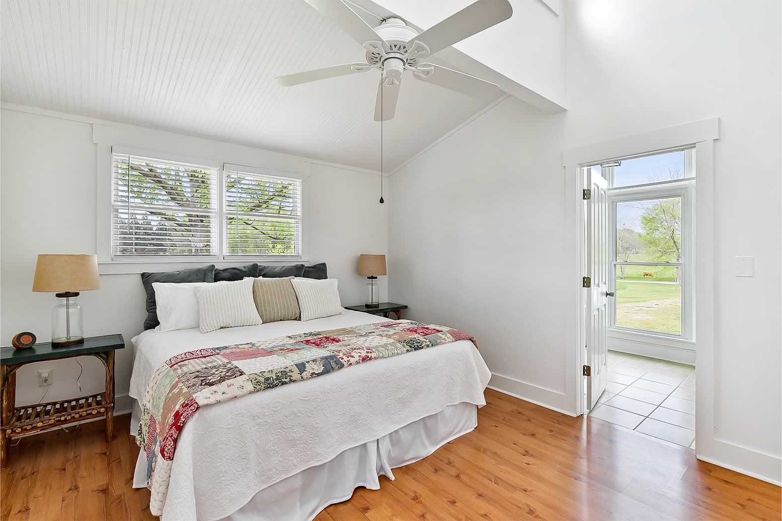 A bright bedroom with vaulted ceilings and wood floors, featuring a king bed with a patchwork quilt and an open door leading to a sunlit, tiled porch with pasture views.