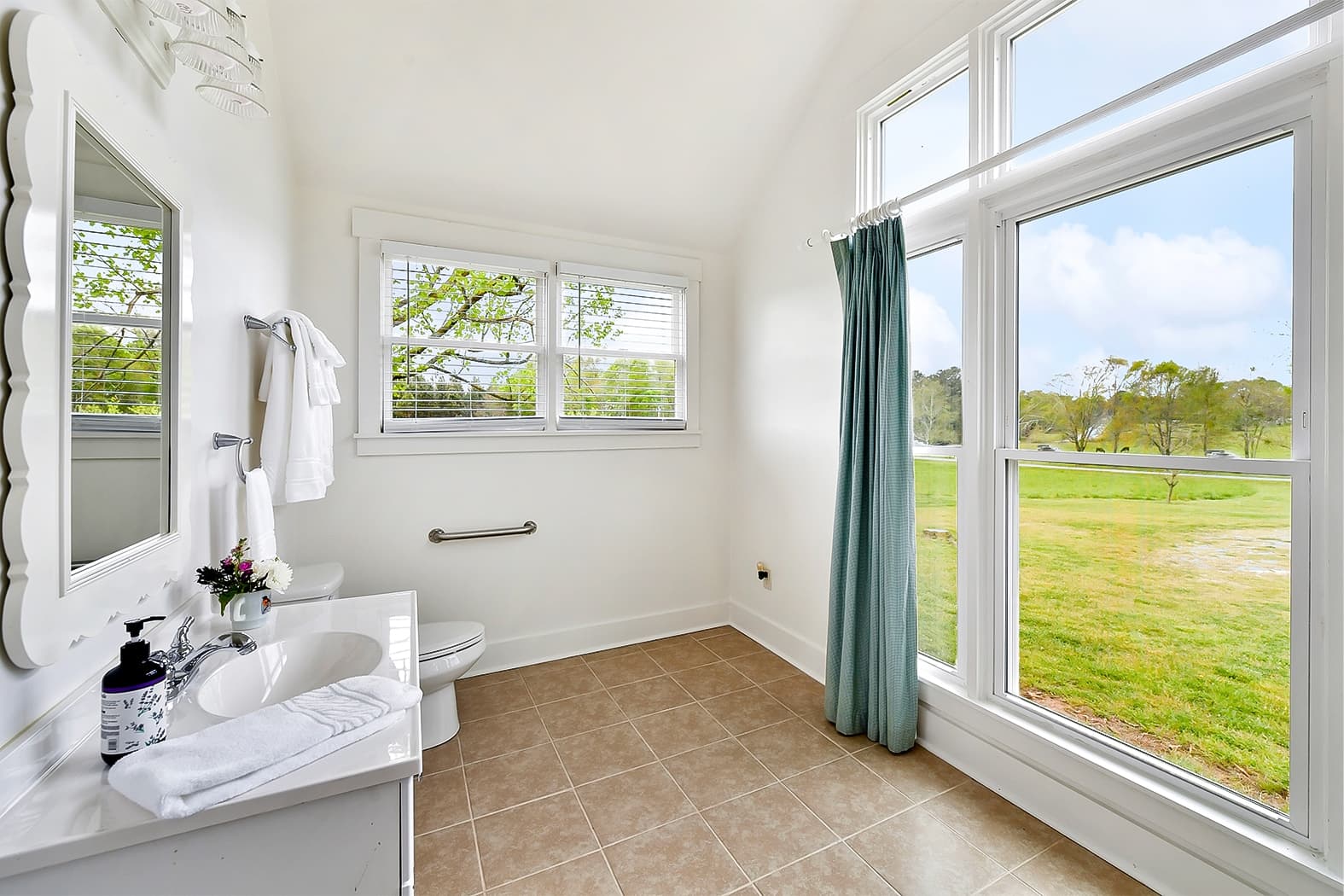 A bright bathroom with tan tile floors and vaulted ceilings, featuring a white vanity and large windows that offer a panoramic view of a green pasture and trees.