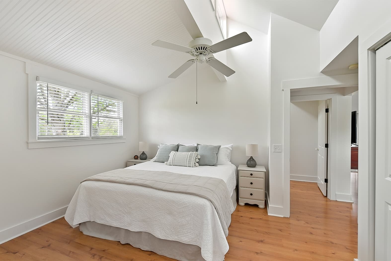 A bright bedroom with vaulted ceilings, a white ceiling fan, and warm wood floors. It features a king bed with white and gray linens, a white nightstand, and a view into a hallway.