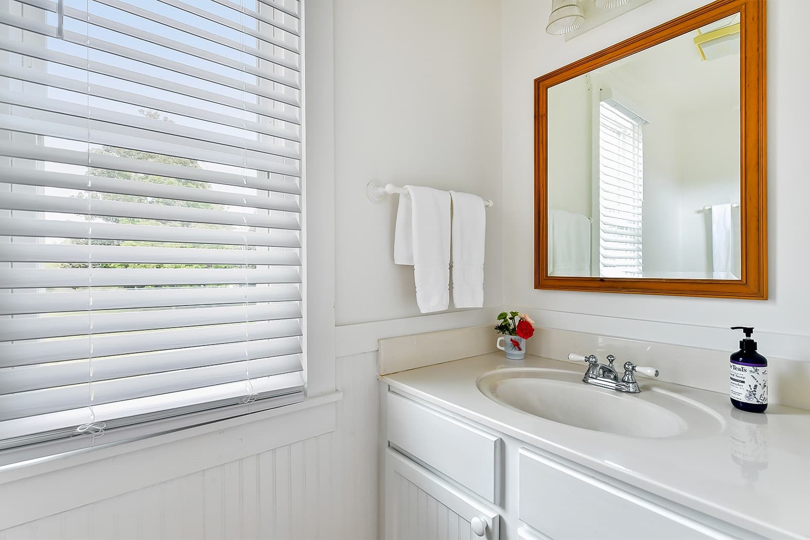 A bright bathroom vanity with a white countertop and sink, a wood-framed mirror, and a window with white blinds that fills the space with natural light.