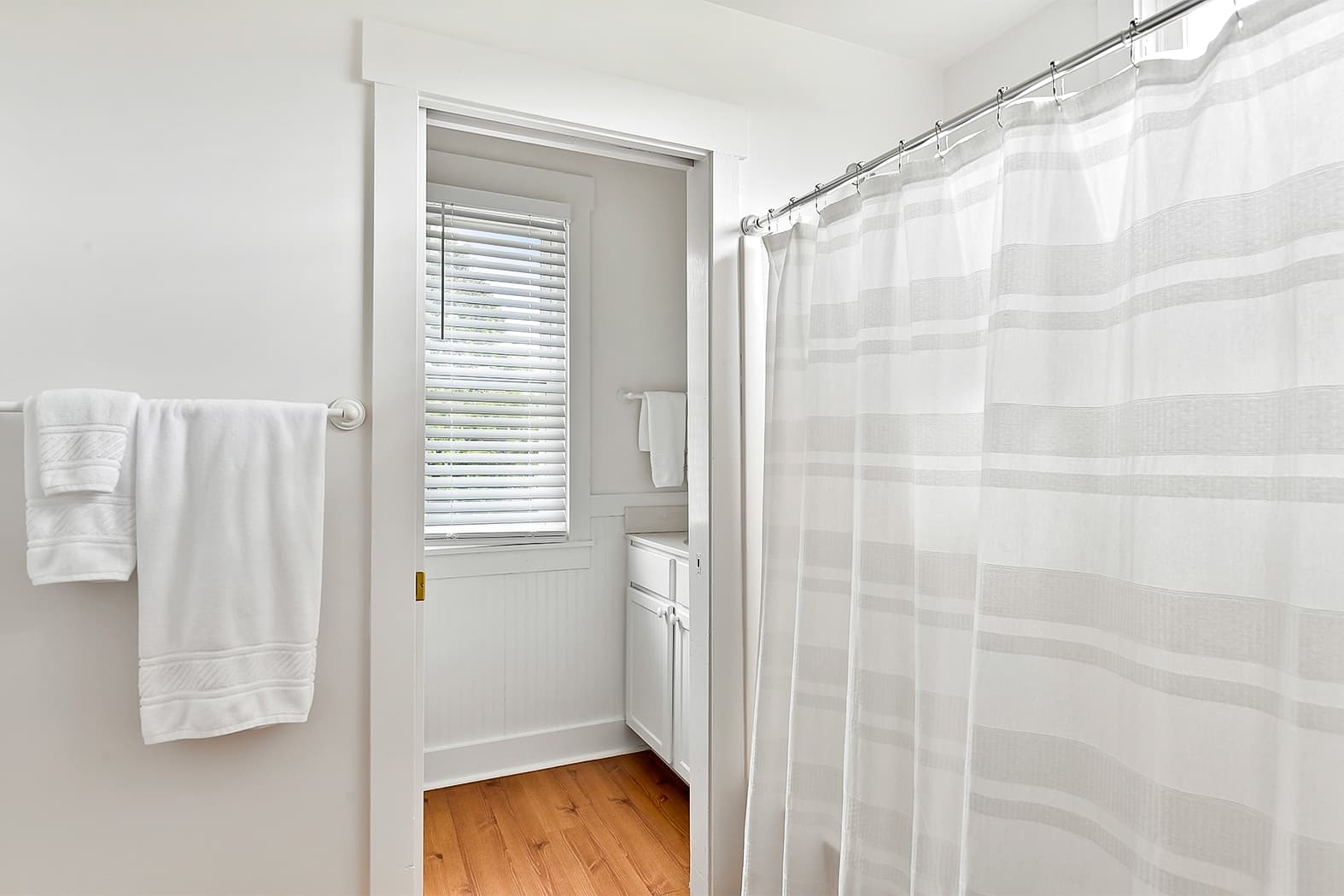 A bright bathroom featuring a white-and-gray striped shower curtain, white towels on a rack, and an open doorway showing a light-filled vanity area with wood floors.