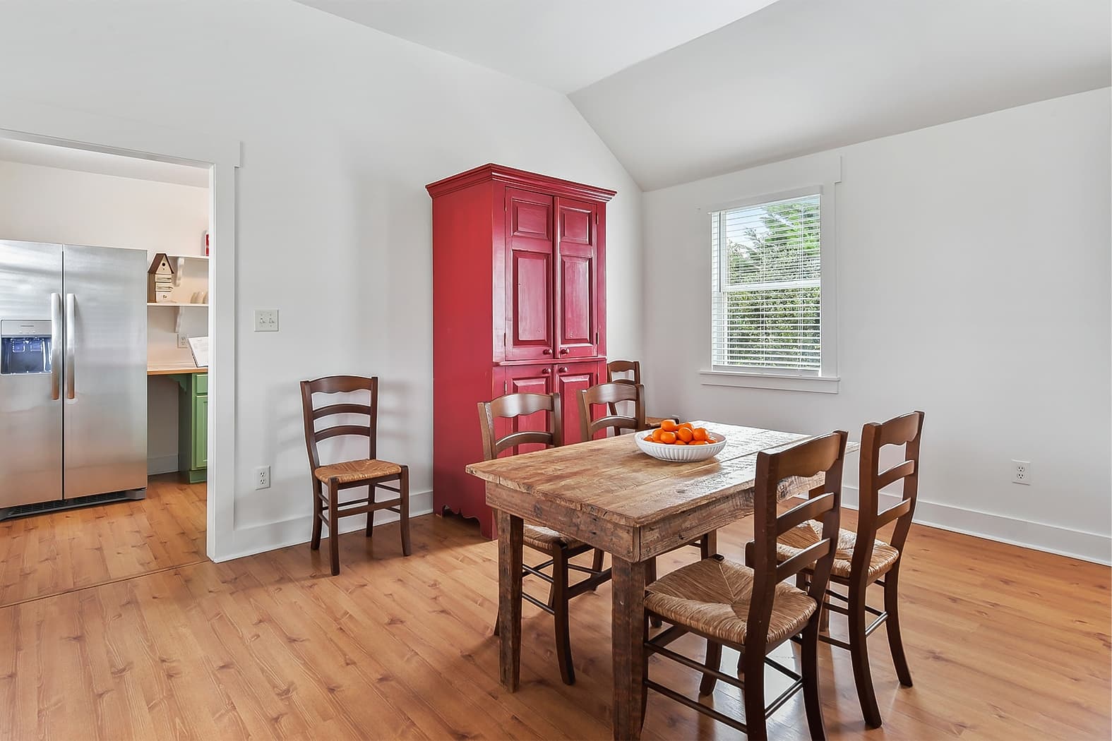 A bright dining area with wood floors and a vaulted ceiling, featuring a rustic wooden table with ladder-back chairs and a bold red armoire. An open doorway provides a view of a modern kitchen with a stainless steel refrigerator.