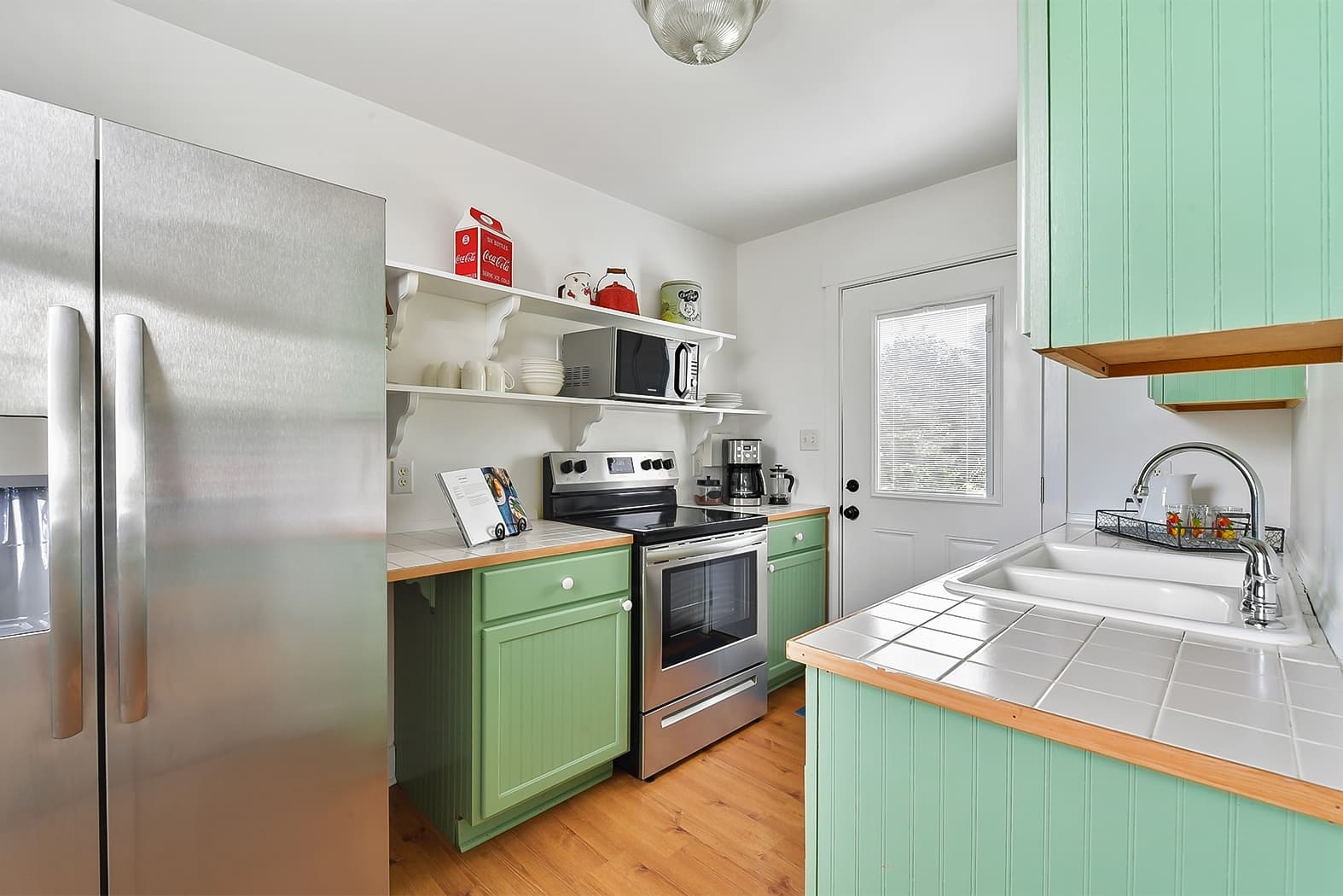 A charming, well-lit kitchen with mint green cabinets, stainless steel appliances, and wood floors. It features white tiled countertops, open shelving with colorful accents, and a double-basin sink.