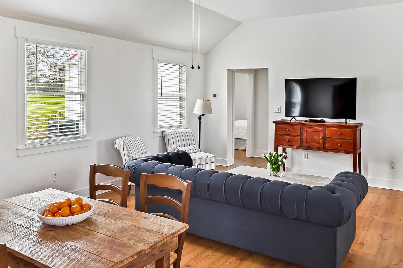A bright, open-concept living area with vaulted ceilings and wood floors, featuring a tufted navy blue sofa, a rustic dining table, and a large flat-screen TV on a wooden console.