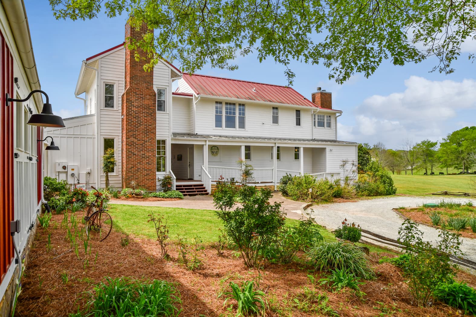An exterior view of a large white farmhouse with a red metal roof and a prominent brick chimney, featuring a long covered porch and landscaped gardens under a blue sky.