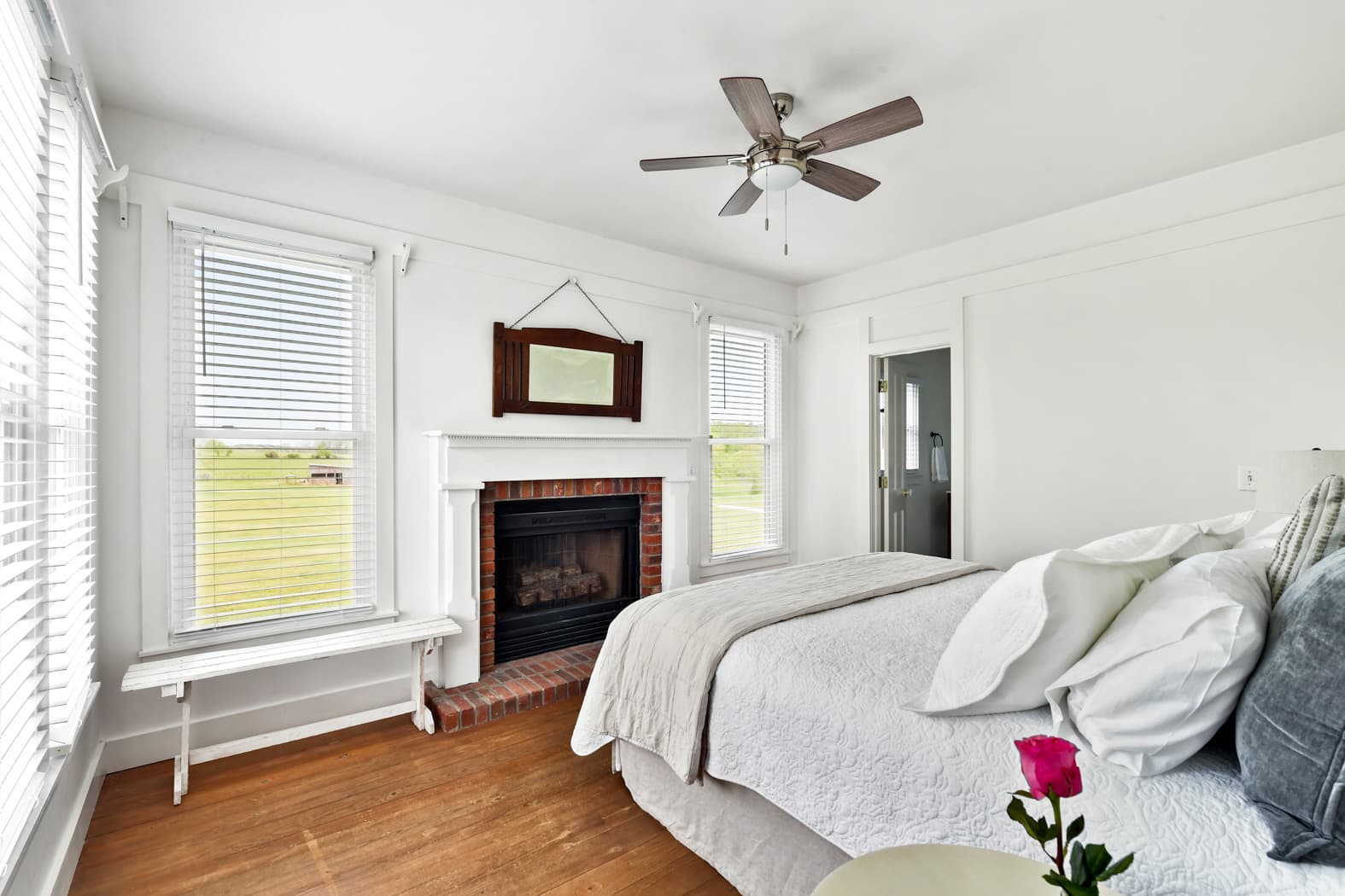 A bright bedroom with wood floors and a ceiling fan, featuring a king bed with white and gray linens, a fireplace with a white mantel, and large windows looking out onto a grassy field.
