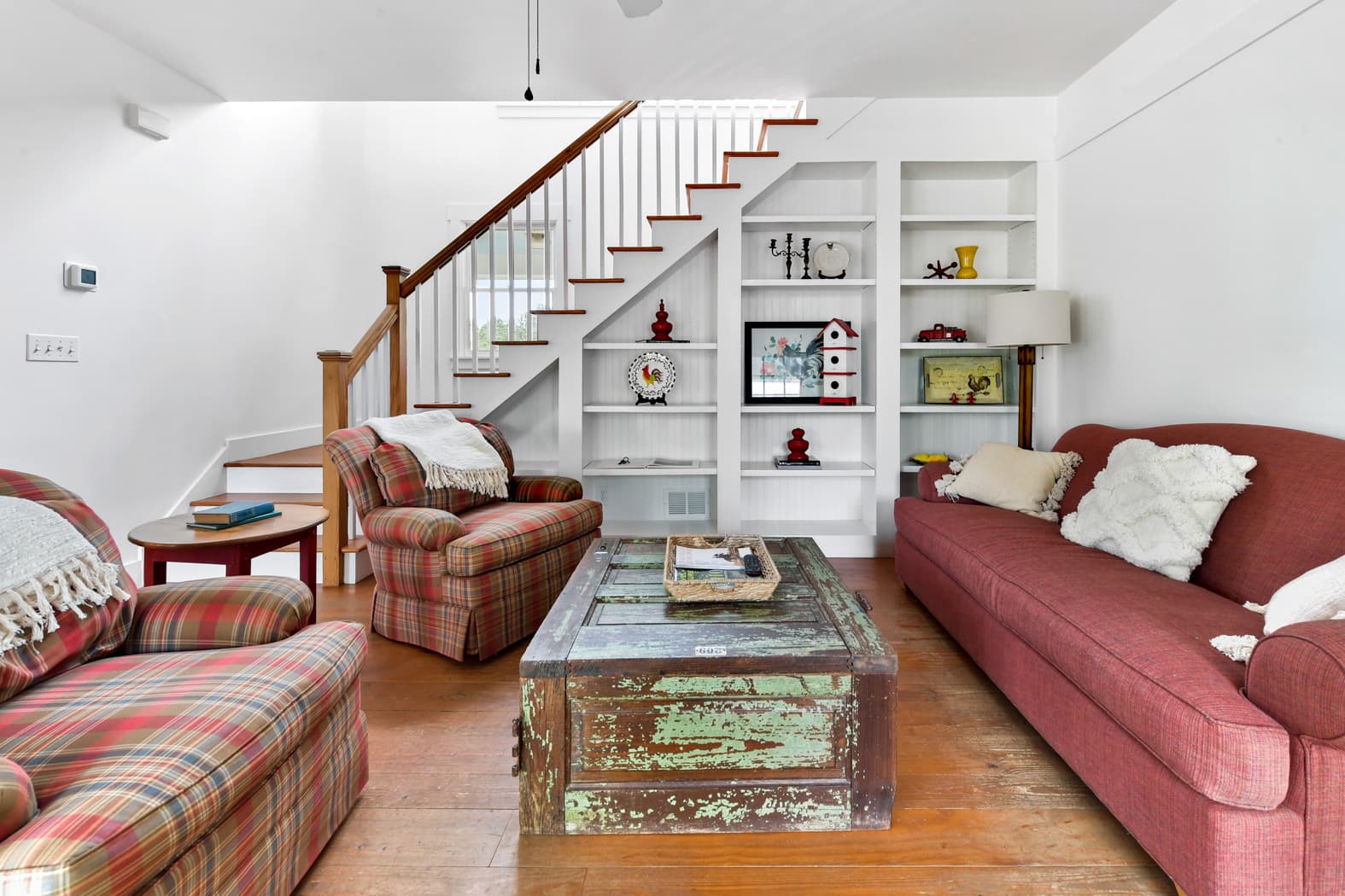 A cozy living area with wood floors and white walls, featuring a red sofa, plaid armchairs, and a rustic trunk used as a coffee table. Built-in white bookshelves are tucked under a wooden staircase.