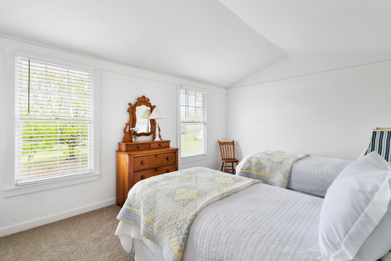 A bright bedroom with white walls and carpet, featuring two twin beds with yellow and gray quilts, a vintage wooden dresser with a mirror, and windows providing a view of green trees.