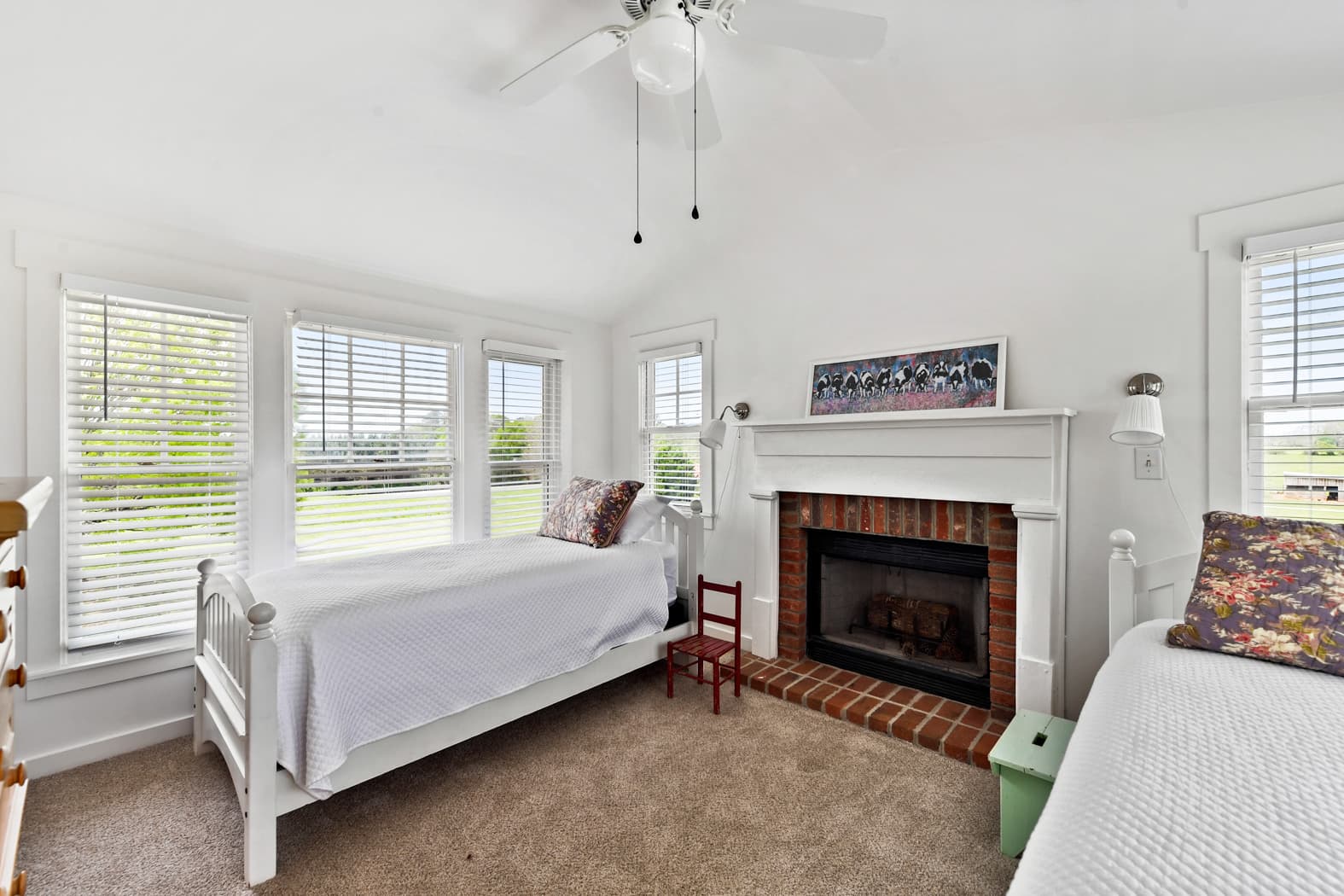 A cozy bedroom with vaulted ceilings and tan carpet, featuring two twin beds with white linens and a white fireplace with a brick hearth. Large windows fill the room with natural light and offer views of the grounds.
