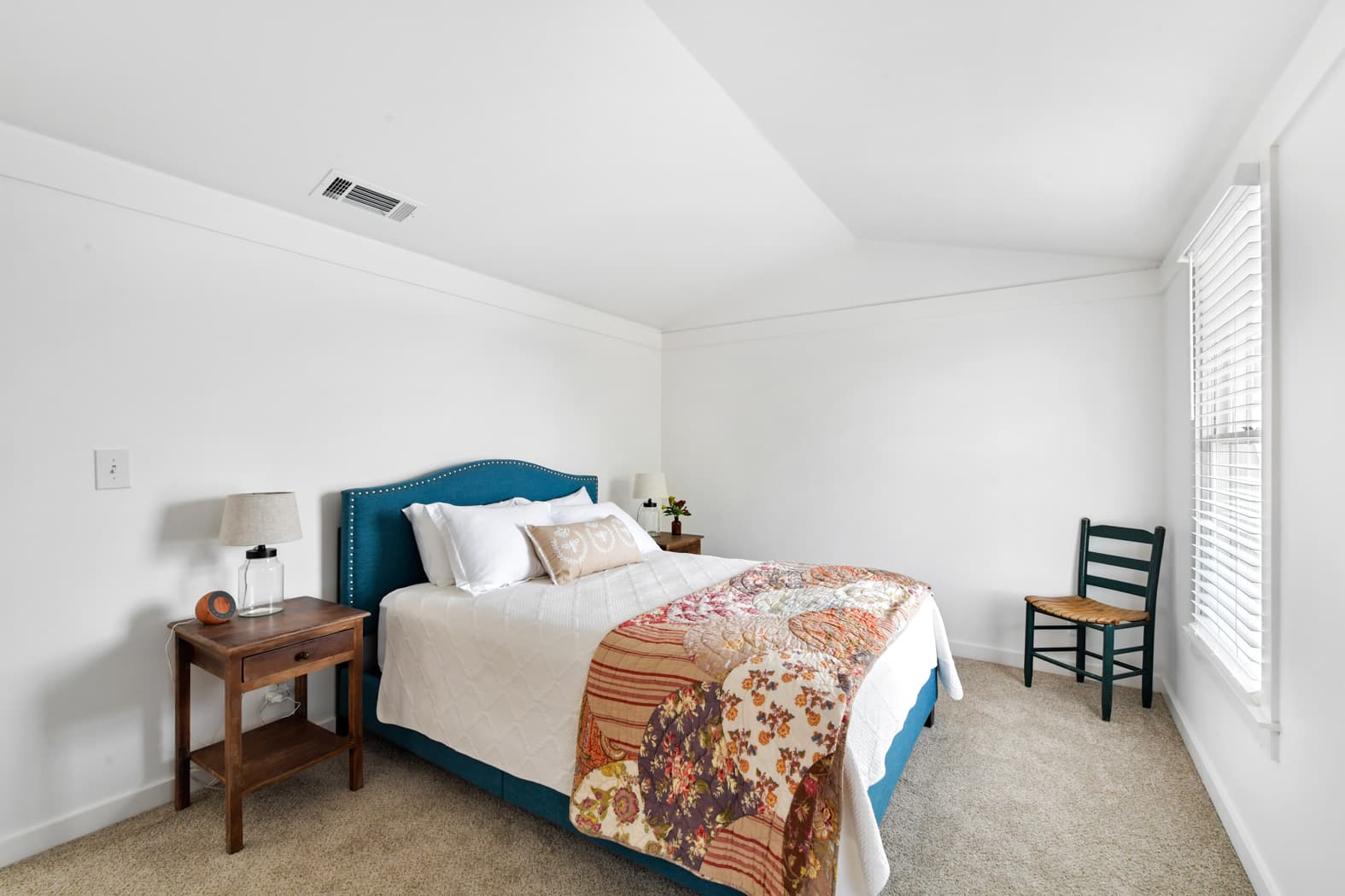 A bright bedroom with vaulted ceilings and tan carpet, featuring a bed with a teal upholstered headboard and a colorful patchwork quilt. A wooden nightstand with a lamp and a teal chair near the window complete the room.