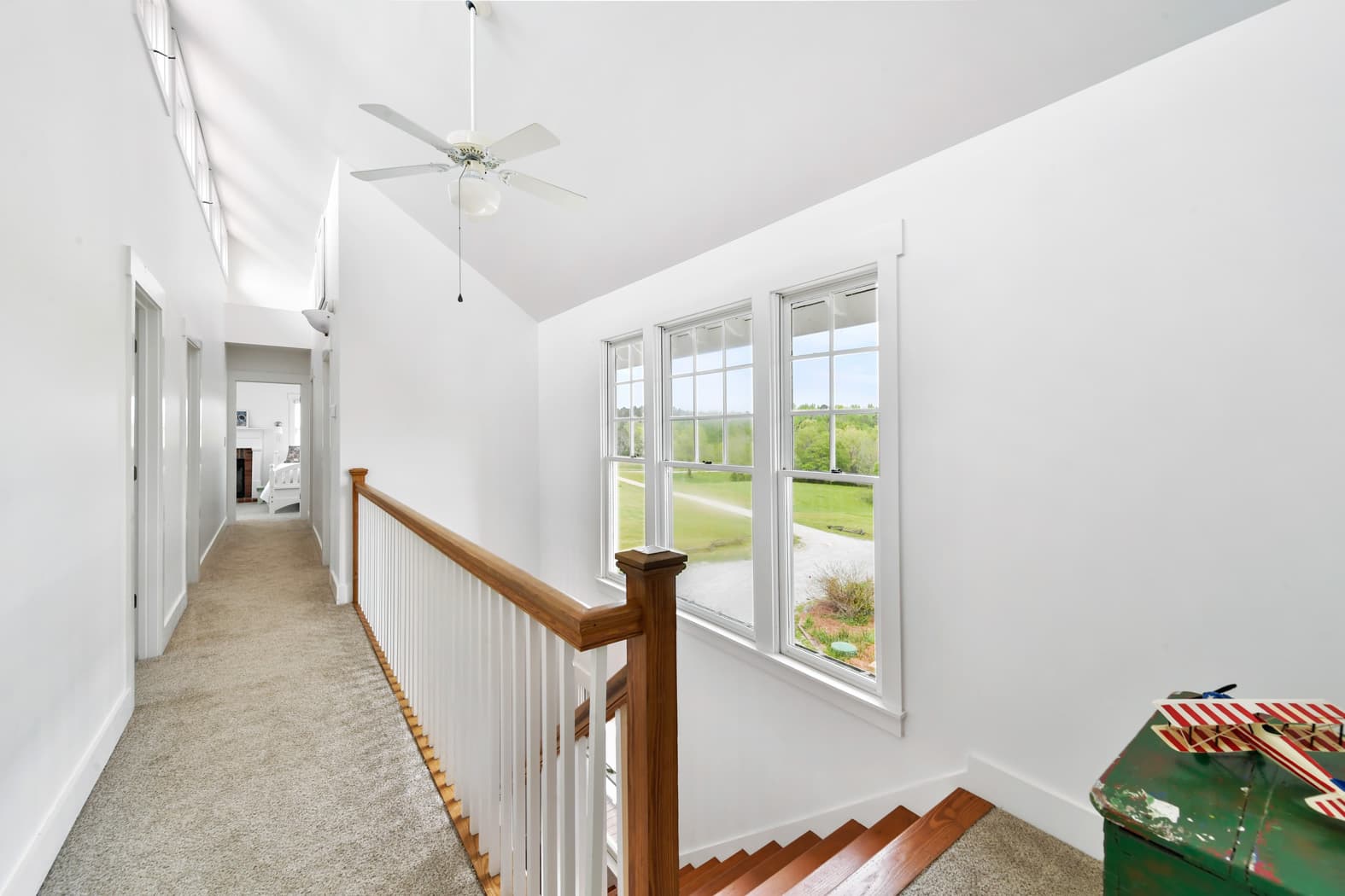 A bright upper-level hallway with vaulted ceilings, tan carpet, and a wooden banister overlooking a staircase. A large window provides a wide view of the green grounds and a winding path.