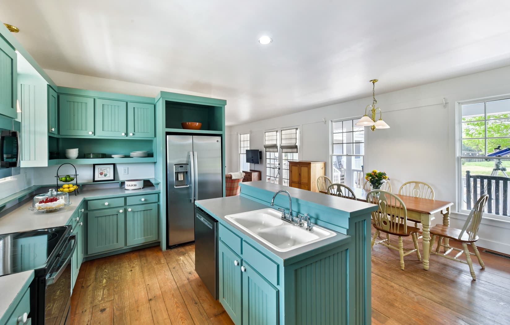 A spacious farmhouse kitchen with teal cabinets and wood floors, featuring a center island with a double sink, stainless steel appliances, and a nearby wooden dining table set for four.