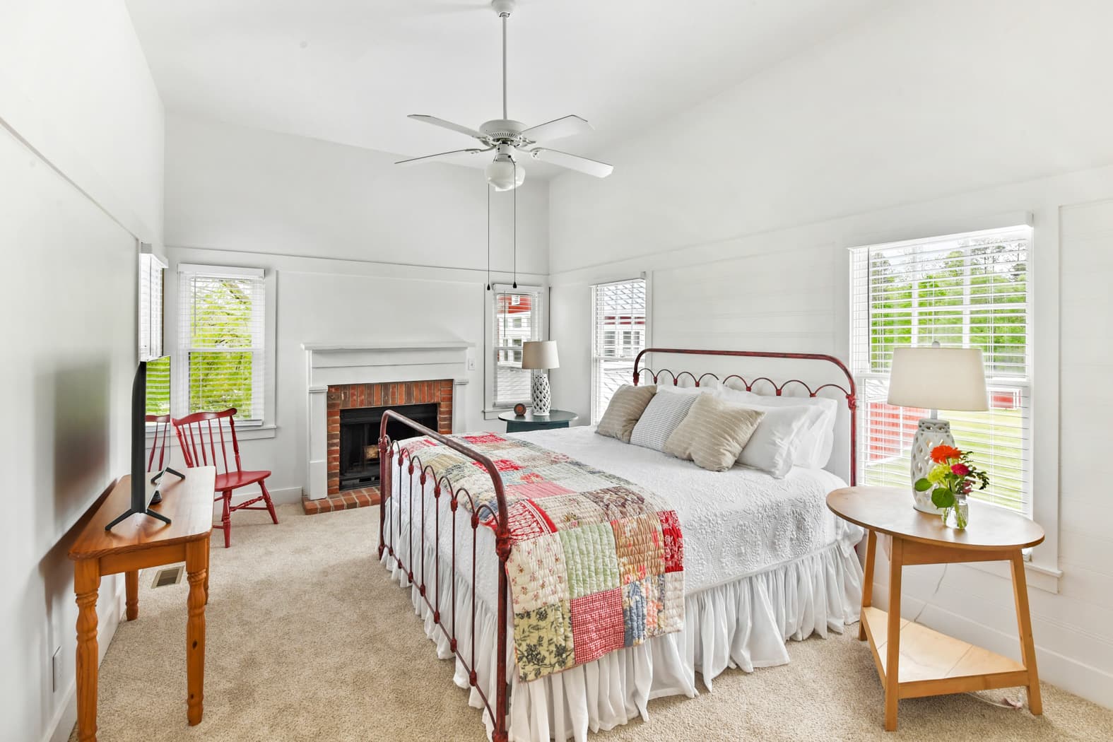 A bright bedroom with white shiplap walls and a vaulted ceiling, featuring a red metal bed frame with a colorful patchwork quilt, a white fireplace with a brick surround, and a wall-mounted TV.