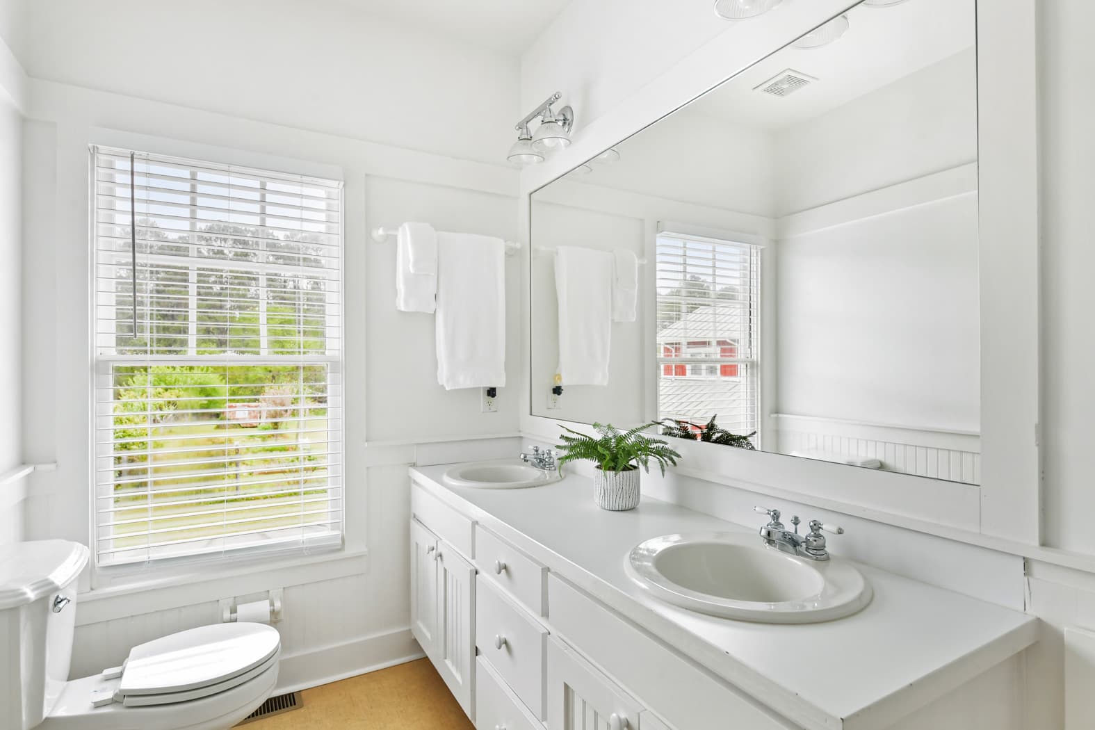 A bright bathroom with a long white double vanity, a large framed mirror, and a window with blinds overlooking a green wooded area. The room features white wall paneling and a clean, airy aesthetic.