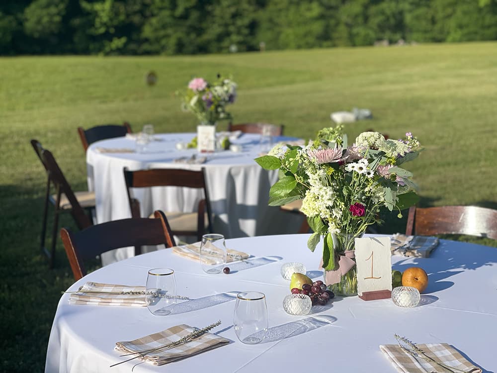 A beautifully set outdoor dining table with a floral centerpiece and elegant tableware, surrounded by green grass.
