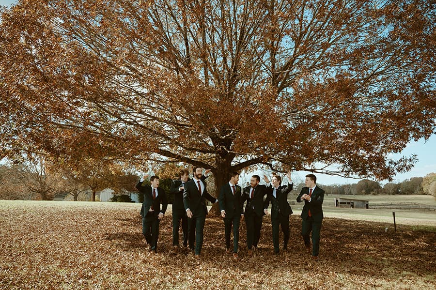 A group of men in suits walks together under a large tree with autumn leaves.