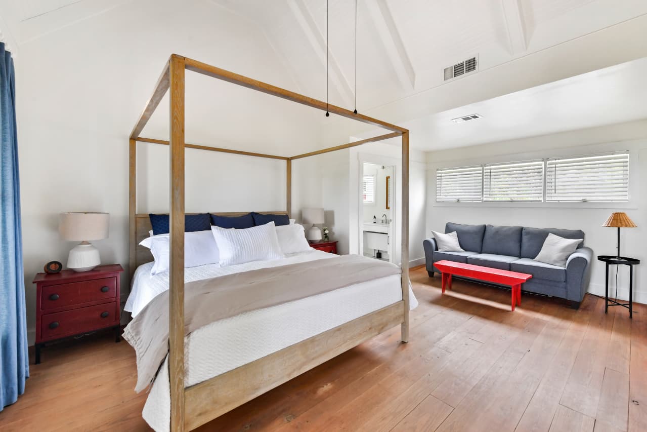 A bright bedroom with vaulted ceilings and wood floors featuring a light wood four-poster bed, a red nightstand, and a blue sofa with a bright red bench in front of it.