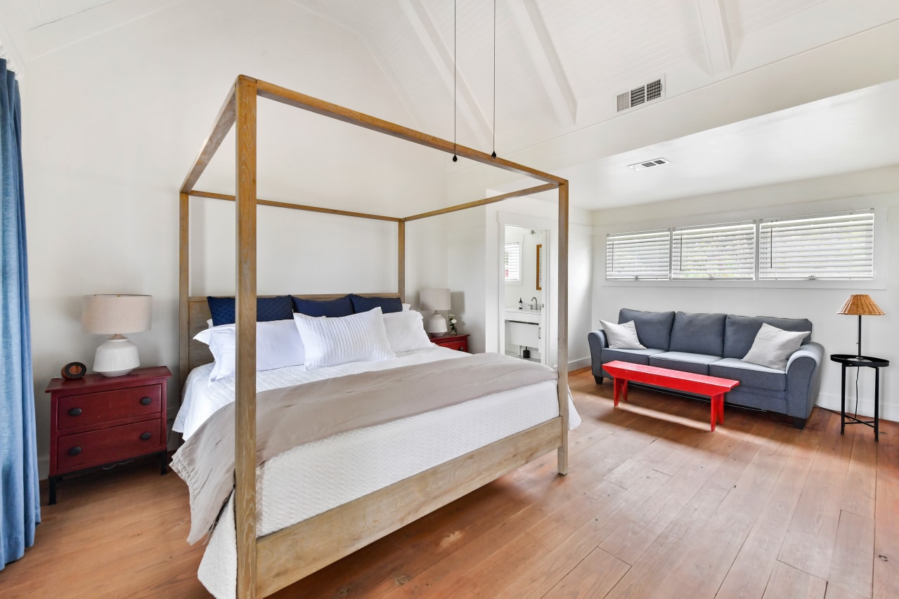 A bright bedroom with vaulted ceilings and wood floors featuring a light wood four-poster bed, a red nightstand, and a blue sofa with a bright red bench in front of it.