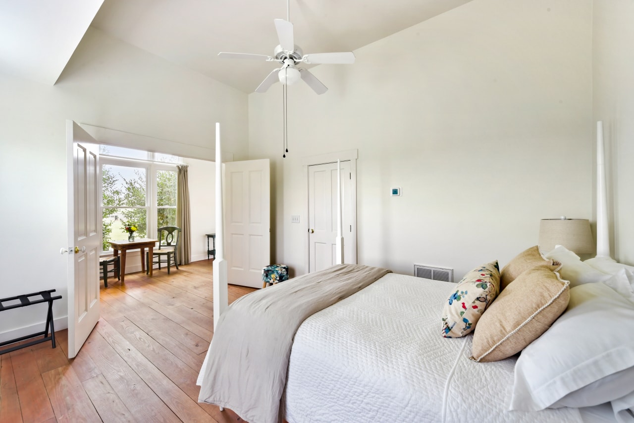 A bright bedroom with vaulted ceilings, wood floors, and a white four-poster bed. An open door leads to a sunlit sitting area with a small wooden table and chairs.