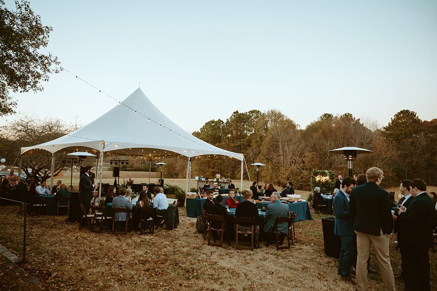 A festive outdoor gathering under a large white tent with guests seated at tables.
