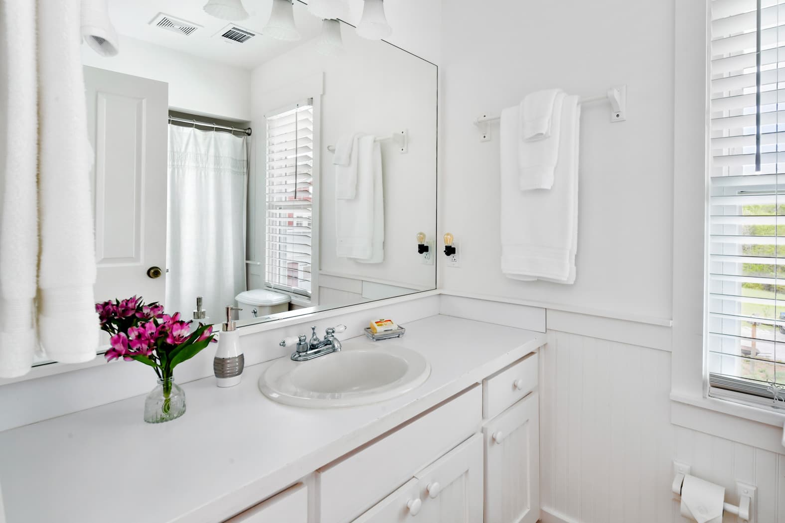 A clean, all-white bathroom vanity with a large frameless mirror, white cabinetry, and a vase of vibrant pink flowers. The reflection shows a white shower curtain and a window with white blinds.