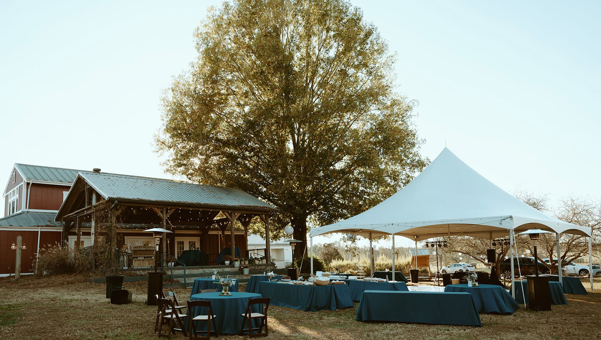 An outdoor event setup featuring tables under a large tent next to a rustic barn and a tall tree.
