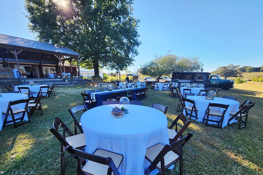 An outdoor event space with round tables set up under a sunny sky, surrounded by greenery.