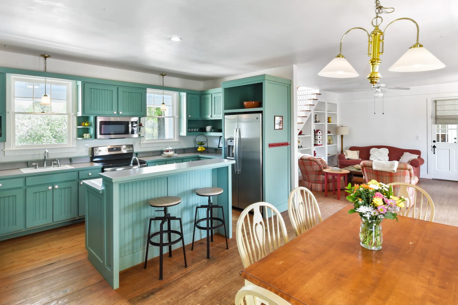 A bright, open-concept farmhouse interior featuring a kitchen with teal cabinetry and a breakfast bar with stools, leading into a dining area and a cozy living room with a red sofa.