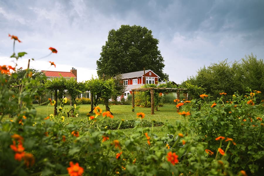 A quaint red house surrounded by vibrant flowers and trees under a cloudy sky.