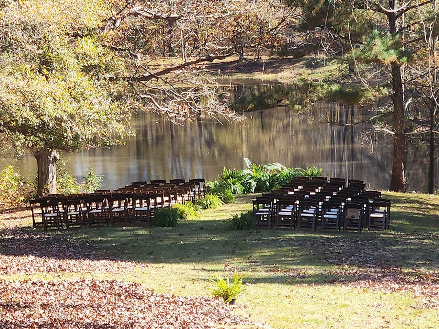 A row of wooden chairs set up for an outdoor event near a tranquil pond surrounded by trees.
