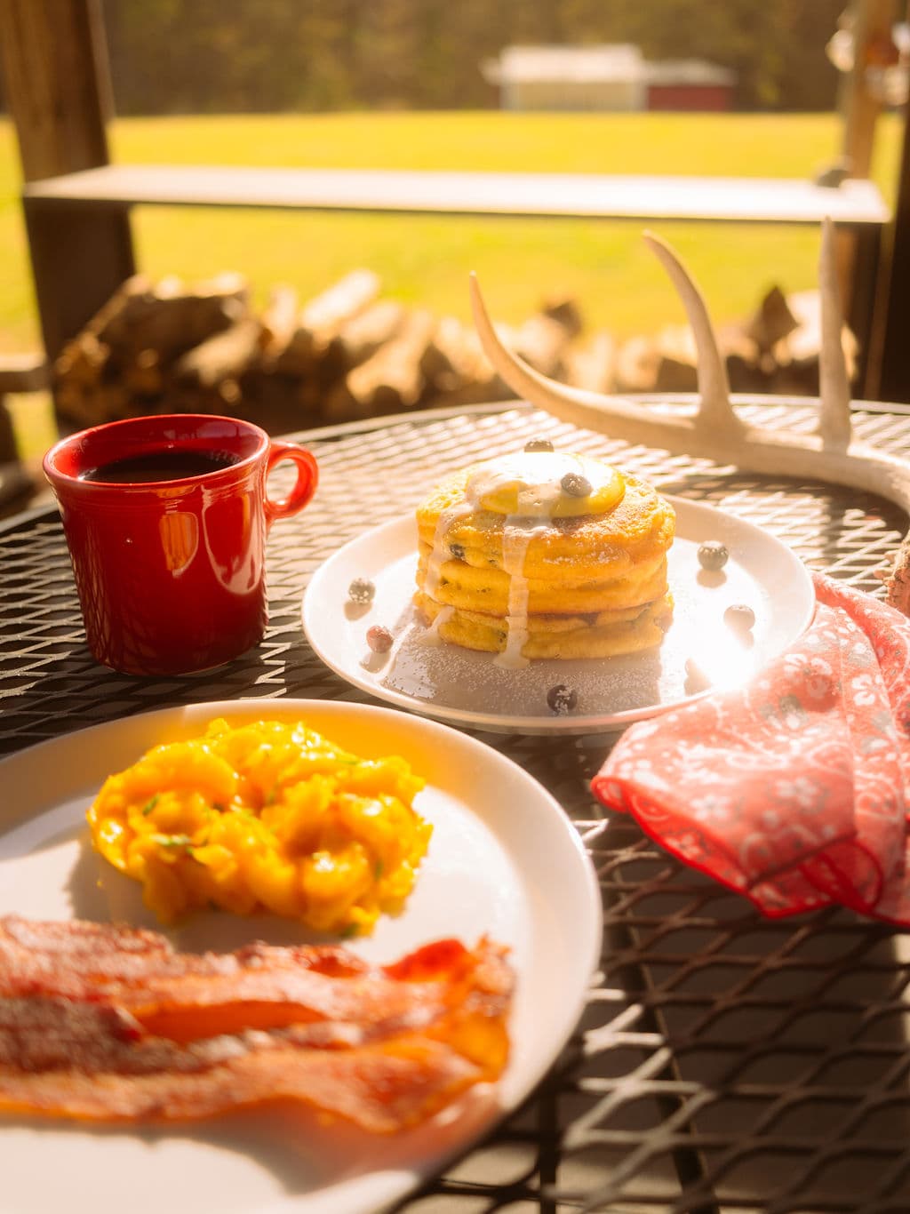 A breakfast spread featuring pancakes with blueberries and syrup, crispy bacon, a side of mango, and a cup of coffee, set on a sunny outdoor table.