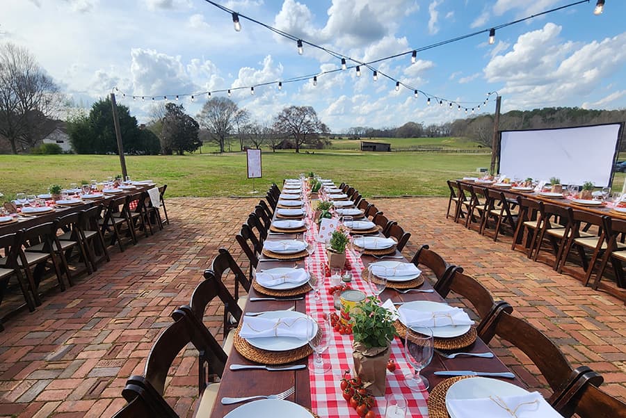 A beautifully set outdoor dining area with long tables adorned with checkered tablecloths, white plates, and greenery, under a clear sky with string lights overhead.